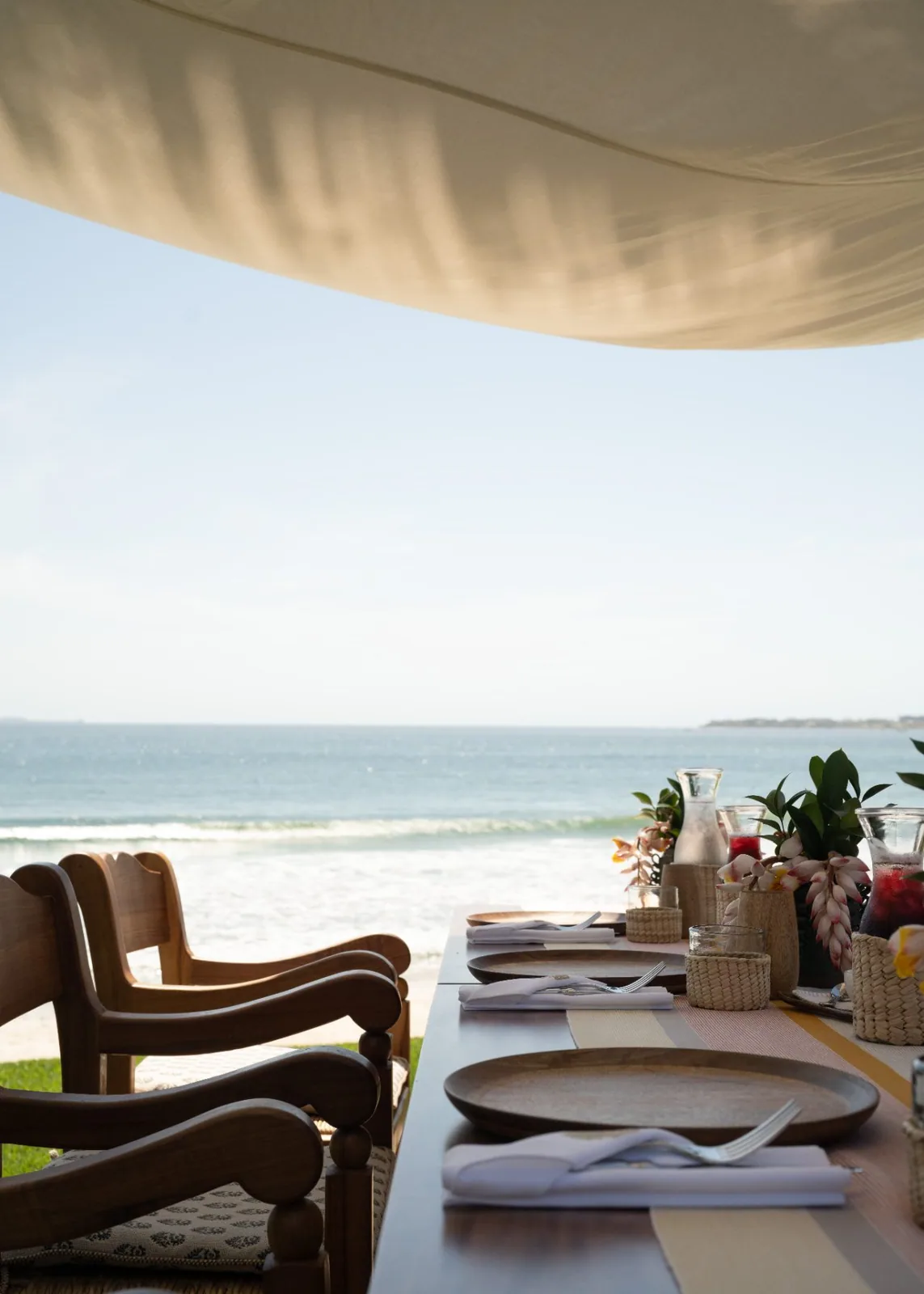 A table laid with linen and rattan chairs on the sand at the water&rsquo;s edge